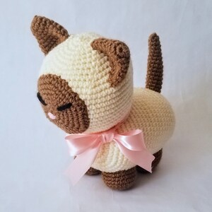May include: A crocheted Siamese cat plush toy with brown ears and a pink bow. The cat is sitting on a white surface.