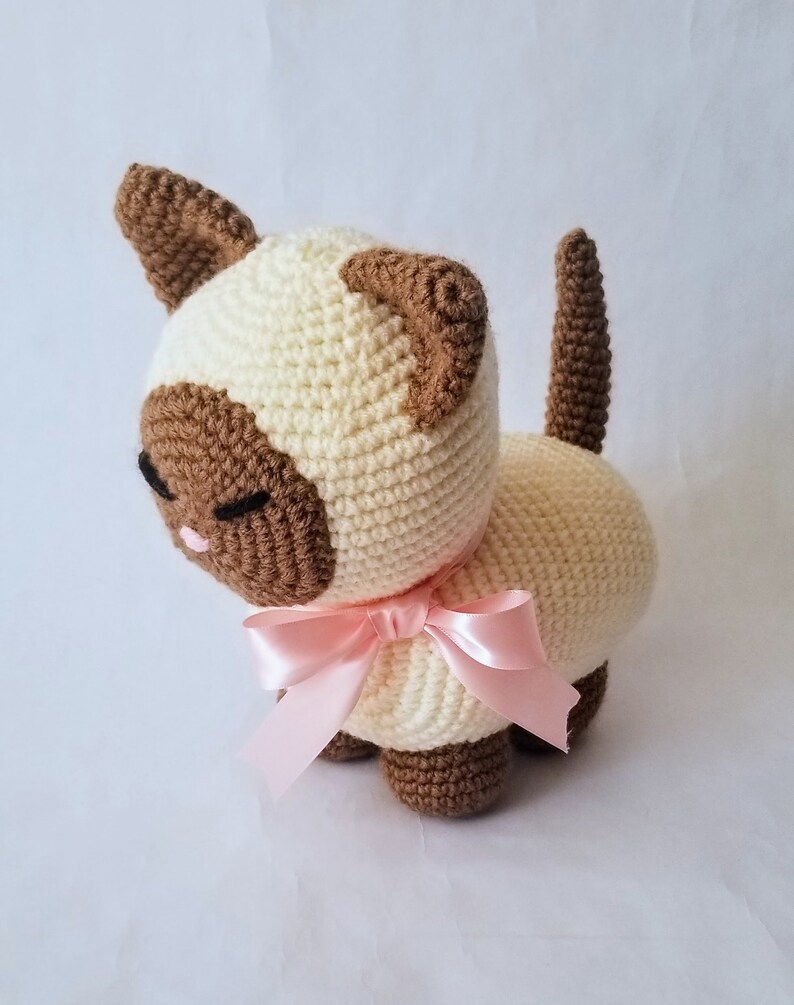 May include: A crocheted Siamese cat plush toy with brown ears and a pink bow. The cat is sitting on a white surface.