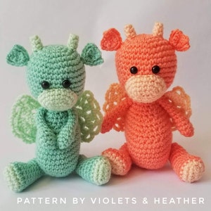 May include: Two crocheted dragon plushies, one green and one orange, sitting on a white surface. They have black eyes and are wearing wings.