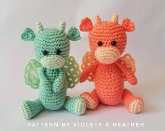 May include: Two crocheted stuffed creatures with small horns, ears, and lacy wings. One is mint green and the other is coral orange. Both have black plastic eyes and a light-colored snout. Text at the bottom reads: PATTERN BY VIOLETS & HEATHER.
