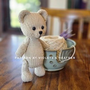 May include: A beige knitted teddy bear with black button eyes and a black nose stands on a wooden surface. The bear is next to a teal ceramic yarn bowl with a ball of yarn and lavender sprigs. Text on the bowl reads "PATTERN BY VIOLETS & HEATHER."