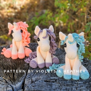May include: Three crocheted unicorn amigurumi toys. The unicorns are sitting on a tree stump. The unicorn on the left has pink yarn for its mane and tail. The unicorn in the middle has purple yarn for its mane and tail. The unicorn on the right has blue yarn for its mane and tail. The text "PATTERN BY VIOLETS & HEATHER" is printed on the tree stump.