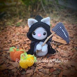 May include: A gray crocheted cat wearing a black hooded cloak and holding a gray crocheted scythe. The cat is sitting on a bed of brown wood chips with two orange and yellow crocheted pumpkins in front of it. The text "Pattern by Violets & Heather" is visible below the cat.