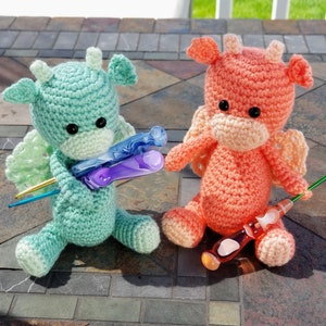 May include: Two crocheted dragon plushies, one green and one peach, holding colorful crochet hooks.