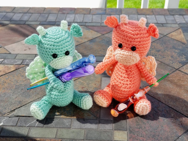 May include: Two crocheted dragon plushies, one green and one peach, holding colorful crochet hooks.