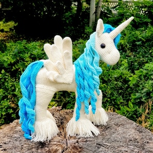 May include: A white crocheted unicorn with a blue mane and tail. The unicorn has a white horn and is standing on a wooden surface.