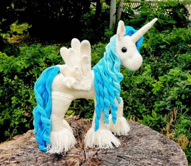 May include: A white crocheted unicorn with a blue mane and tail. The unicorn has a white horn and is standing on a wooden surface.