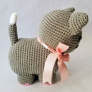 May include: A gray crocheted cat toy with a pink satin ribbon tied around its neck. The cat has a white crocheted tail.