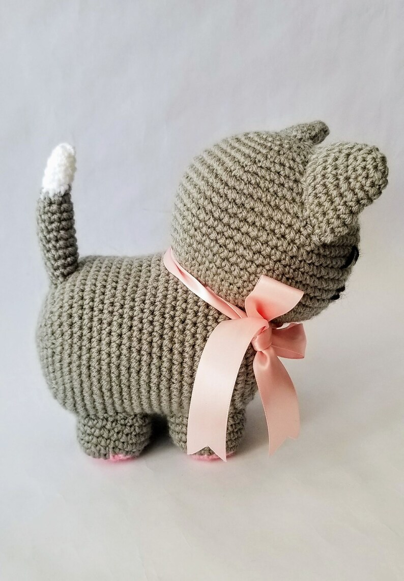 May include: A gray crocheted cat toy with a pink satin ribbon tied around its neck. The cat has a white crocheted tail.