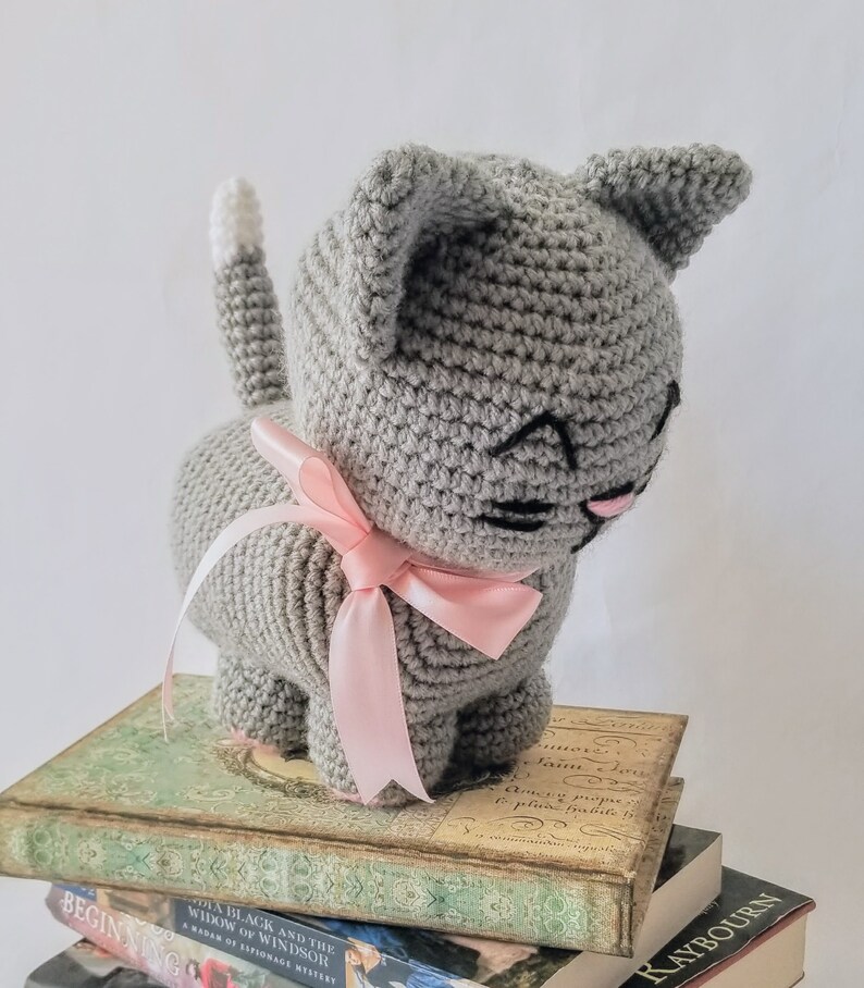 May include: A gray crocheted cat toy with a pink bow. The cat is sitting on a stack of books.