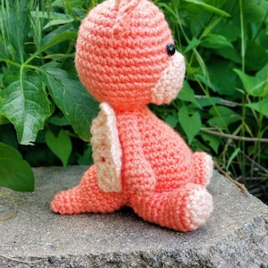 May include: A crocheted pink dragon with white wings sitting on a gray stone. The dragon has black eyes and a pink nose.
