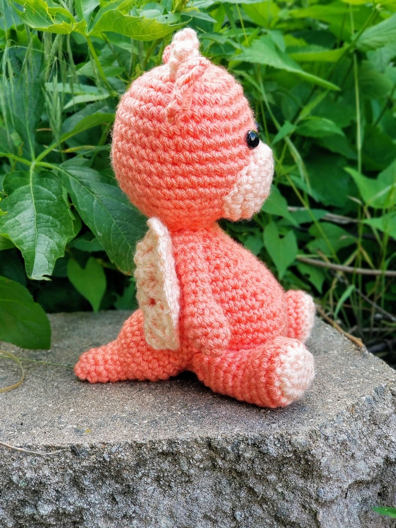 May include: A crocheted pink dragon with white wings sitting on a gray stone. The dragon has black eyes and a pink nose.