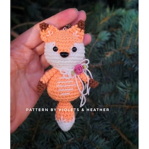 May include: A crocheted orange fox with white accents and a pink flower. The fox has a small, white tail and black eyes. The fox is holding a white ribbon with a pink flower.