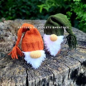 May include: Two knitted gnomes, one with an orange hat and the other with a green hat, sitting on a tree stump. The gnomes have white beards and are wearing knitted hats with tassels. The text "PATTERN BY VIOLETS & HEATHER" is visible on the image.