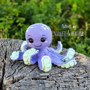 May include: A purple knitted octopus with green tentacles and white spots. The octopus has a smiling face with black eyes and pink cheeks. The octopus is sitting on a wooden surface. Pattern by Violets & Heather.