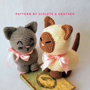 May include: Two crocheted cat toys, one gray and one white with brown markings. Both cats have pink bows around their necks and are sitting on a vintage book.