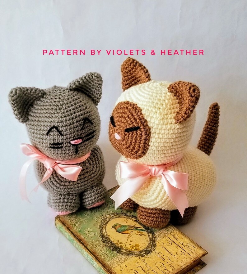 May include: Two crocheted cat toys, one gray and one white with brown markings. Both cats have pink bows around their necks and are sitting on a vintage book.