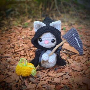 May include: A gray crocheted cat wearing a black hooded cloak and holding a gray crocheted scythe. The cat is standing on a bed of brown wood chips. A small yellow crocheted pumpkin sits in front of the cat.
