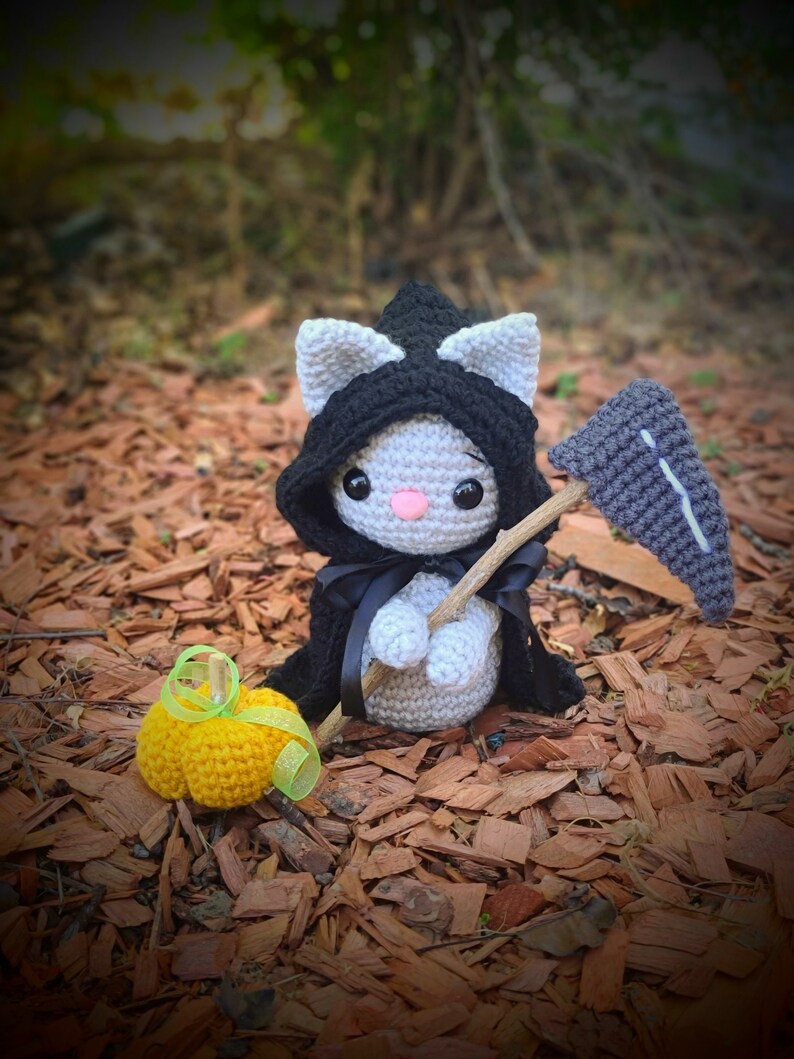 May include: A gray crocheted cat wearing a black hooded cloak and holding a gray crocheted scythe. The cat is standing on a bed of brown wood chips. A small yellow crocheted pumpkin sits in front of the cat.
