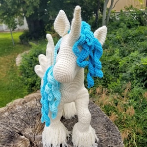 May include: A white crocheted unicorn with a blue mane and tail. The unicorn is standing on a tree stump.