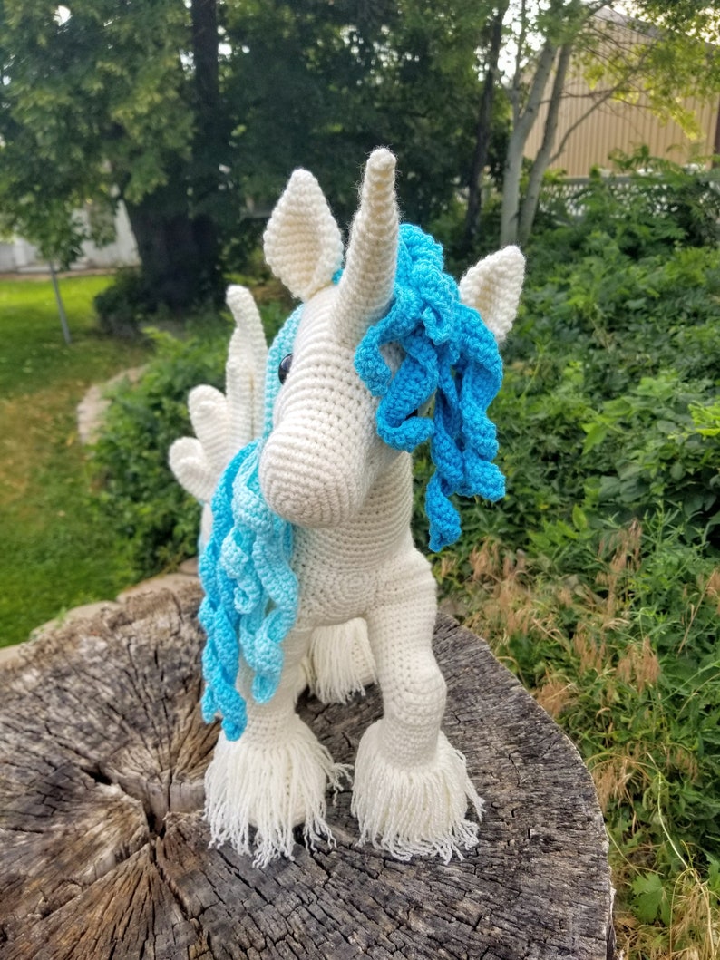 May include: A white crocheted unicorn with a blue mane and tail. The unicorn is standing on a tree stump.