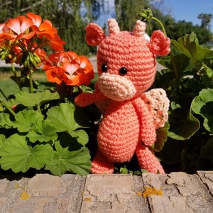 May include: A crocheted pink dragon toy with a light pink nose and black eyes. The dragon is sitting on a brick wall with green plants in the background.
