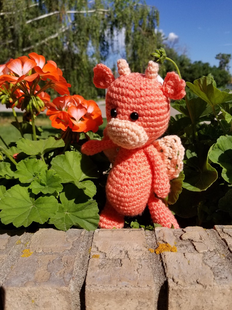 May include: A crocheted pink dragon toy with a light pink nose and black eyes. The dragon is sitting on a brick wall with green plants in the background.