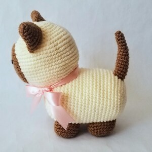 May include: A crocheted cream-colored cat with brown ears and tail, wearing a pink ribbon around its neck.