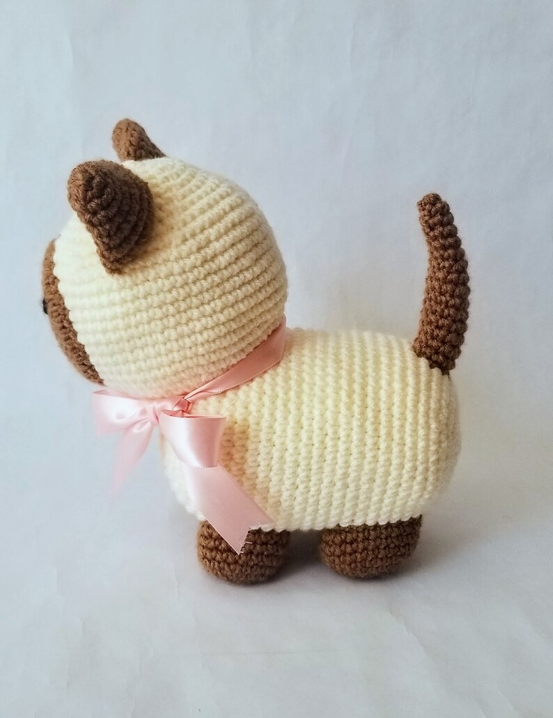 May include: A crocheted cream-colored cat with brown ears and tail, wearing a pink ribbon around its neck.