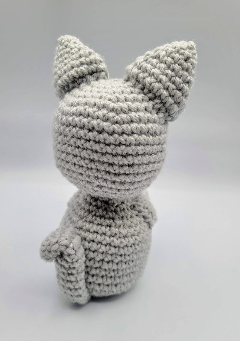 May include: A gray crocheted cat toy with large ears. The cat is sitting on a white surface.