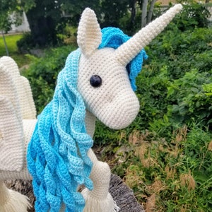 May include: A white crocheted unicorn with a blue mane and horn. The unicorn is standing on a tree stump.