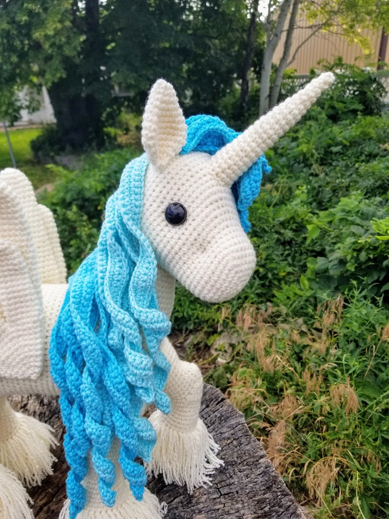 May include: A white crocheted unicorn with a blue mane and horn. The unicorn is standing on a tree stump.