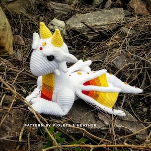 May include: White crocheted dragon with yellow and orange accents. The dragon has a long tail and a small, pointed snout. The dragon is lying on a bed of brown twigs and rocks. "PATTERN BY VIOLETS & HEATHER" is written on the bottom of the image.