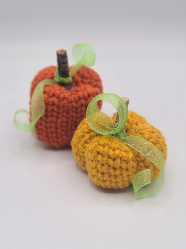 May include: Two knitted pumpkins, one orange and one yellow, with green ribbon accents.