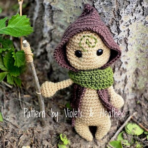 Puede incluir: Muñeca de ganchillo con una capa con capucha marrón, una bufanda verde y un pequeño bastón de madera. La muñeca tiene ojos negros y un diseño bordado verde en la cabeza. El texto "Pattern by Violets & Heather" es visible.
