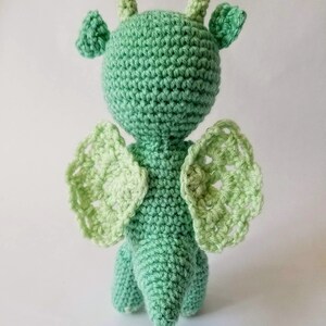 May include: A green crocheted dragon toy with light green wings. The dragon has a long tail and a small head with two horns.