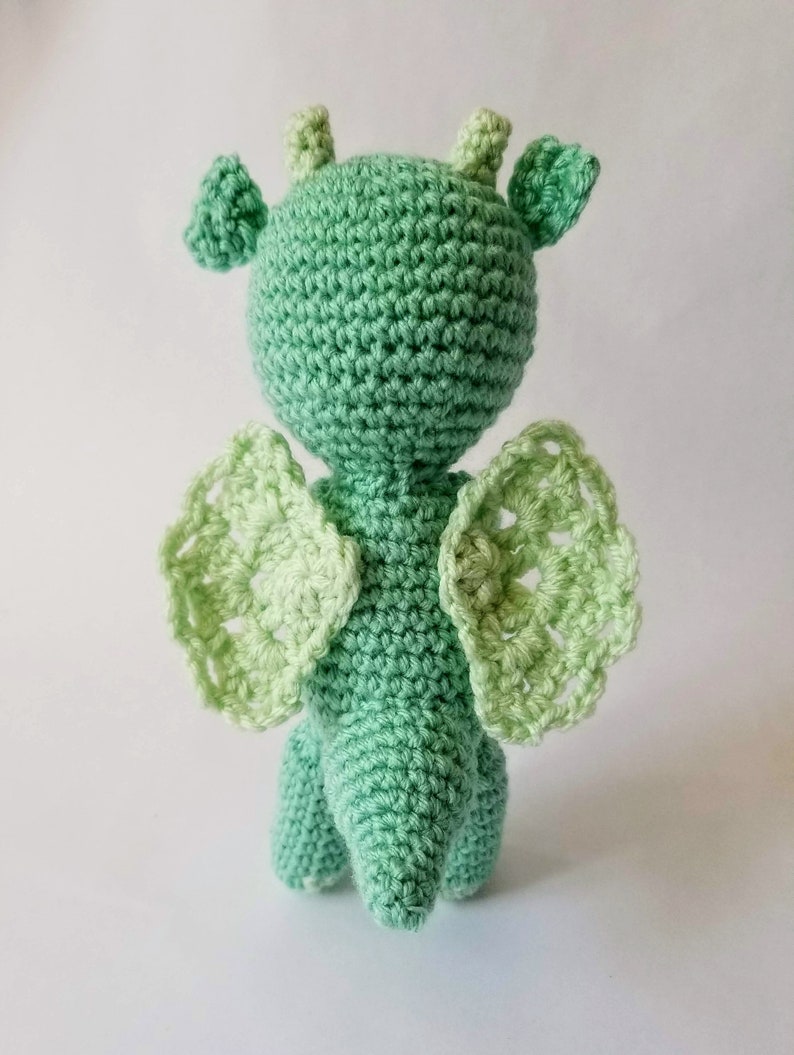 May include: A green crocheted dragon toy with light green wings. The dragon has a long tail and a small head with two horns.