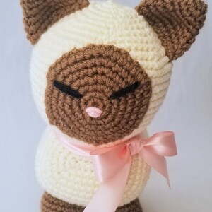 May include: A crocheted stuffed cat toy with brown ears, a brown face, and a pink nose. The cat is wearing a pink satin ribbon around its neck.