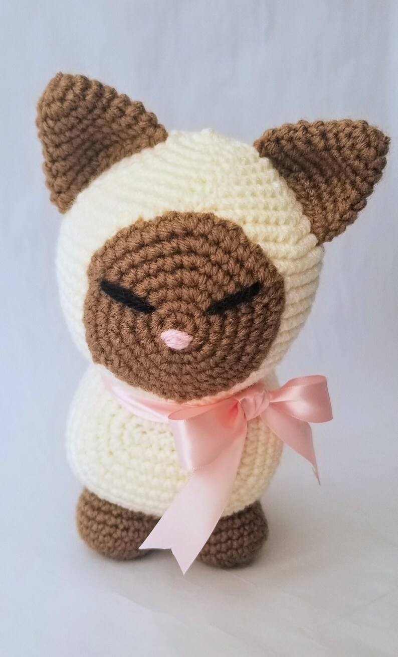 May include: A crocheted stuffed cat toy with brown ears, a brown face, and a pink nose. The cat is wearing a pink satin ribbon around its neck.