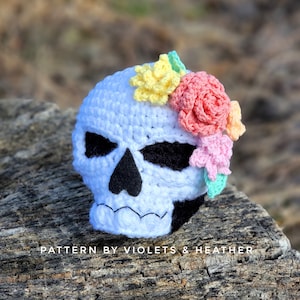 Könnte beinhalten: Weißer gehäkelter Schädel mit schwarzen Augen und einem Mund, verziert mit einer Blumenkrone aus rosa, gelben und grünen Blumen. Der Schädel sitzt auf einem Stück verwittertem Holz. "PATTERN BY VIOLETS & HEATHER" steht auf dem Holz.