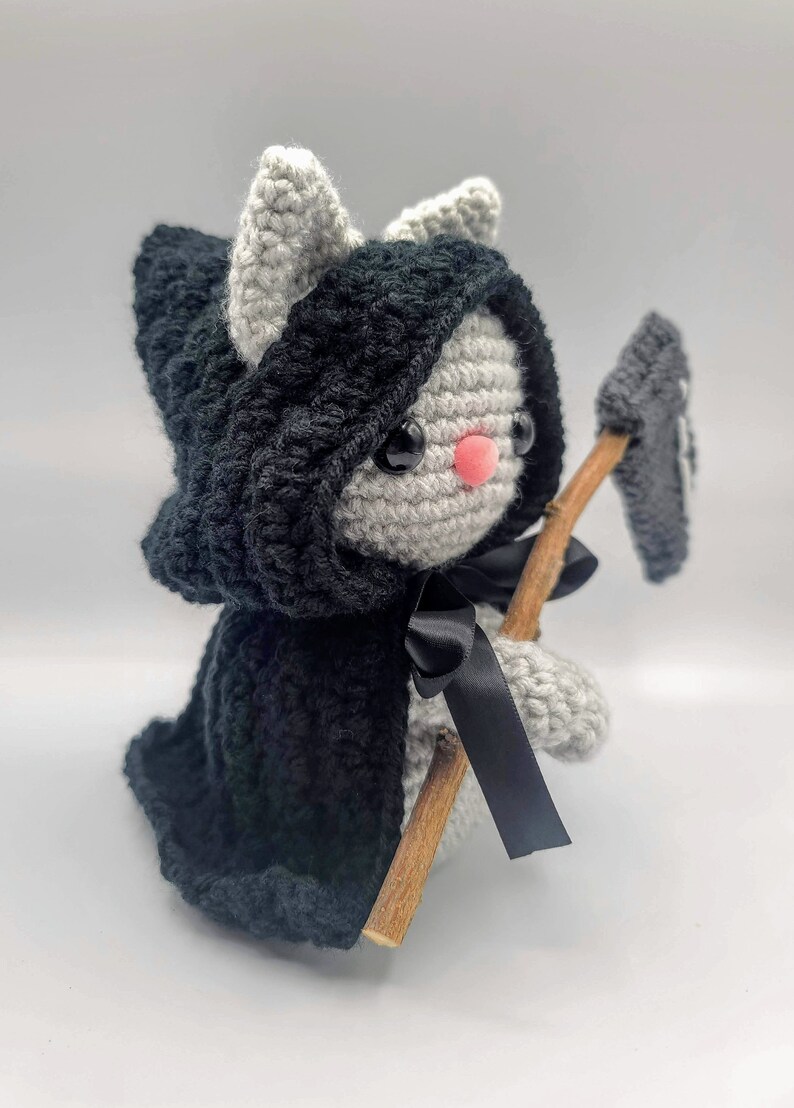 May include: A gray crocheted cat wearing a black hooded cape and holding a wooden stick with a black crocheted scythe.