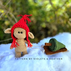 以下が含まれることがあります： 赤い帽子をかぶった編みぐるみのノームが、茶色のそりに載せられた小さな緑色の編みぐるみのクリスマスツリーを引いています。ノームとそりは白い雪の表面にあります。ノームの下には「PATTERN BY VIOLETS & HEATHER」と書かれています。