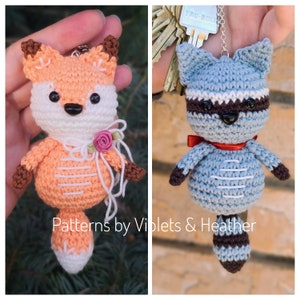 May include: Two crocheted amigurumi keychains, one orange fox and one gray raccoon, both with white accents and black eyes. The fox has a pink flower on its neck. The raccoon has a red bow. The keychains are hanging from a silver chain.