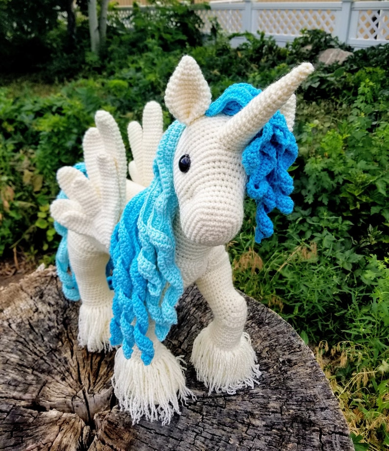 May include: A white crocheted unicorn with blue and light blue yarn mane and tail. The unicorn has a white horn and wings.