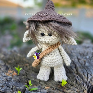 Puede incluir: Un gnomo de crochet que lleva un sombrero marrón y lleva una pequeña bolsa con flores moradas. El gnomo está de pie sobre un trozo de corteza de árbol con plantas verdes que salen de él. El texto "PATTERN BY VIOLETS & HEATHER" es visible en la imagen.