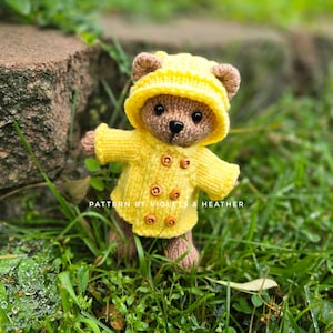 Op de afbeelding: Een gebreide teddybeer met een gele jas met capuchon en houten knopen, staand in het gras. De beer heeft bruine vacht. De tekst "PATTERN BY VIOLETS & HEATHER" is zichtbaar.