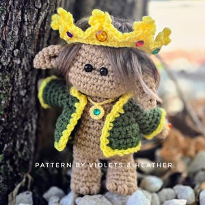 May include: A crocheted elf doll wearing a yellow crown with colourful gems, a green jacket with yellow trim, and a green necklace with a gem. The doll is standing on a bed of white stones. "PATTERN BY VIOLETS & HEATHER" is written below the doll.