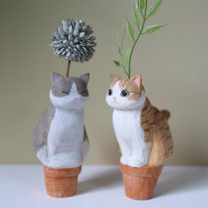 May include: Two hand-carved wooden cat figurines, each perched atop a small terracotta-colored pot. The left cat is grey and white with closed eyes and a grey flower, while the right cat is orange and white with large eyes and a green leaf.
