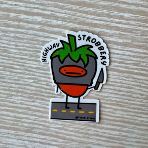 May include: A sticker featuring a cartoon strawberry character with a gray head, orange body, and green leaves. The strawberry holds a knife and stands on a road. Text reads "HIGHWAY STROBBERY" and "@TYPER_STUDIO".