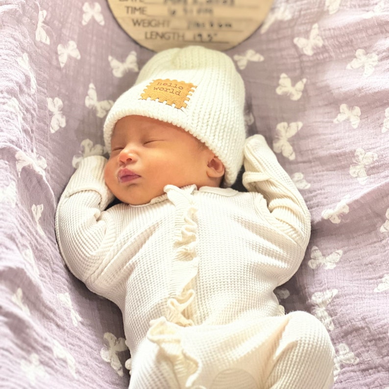 Pu&ograve; includere: Un neonato addormentato indossa un cappello di maglia color crema con una toppa "hello world" e una tutina a nido d'ape abbinata. Il bambino &egrave; su una coperta lavanda con farfalle. La lunghezza del bambino &egrave; di 49,5 cm.